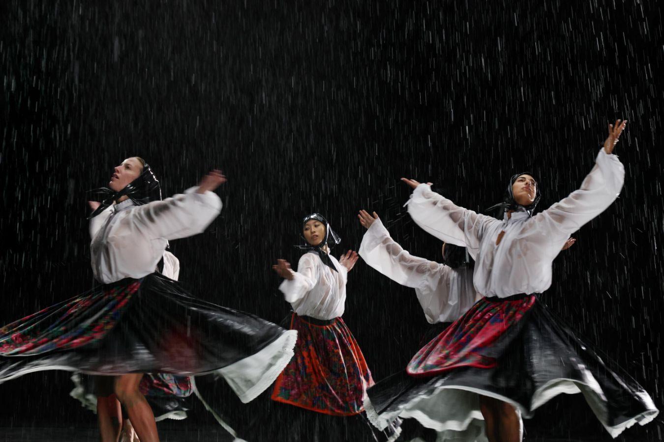 Four people perceived as female wearing black headscarves, white blouses, and black skirts raise their arms in the air. The background is black and raindrops can be seen.