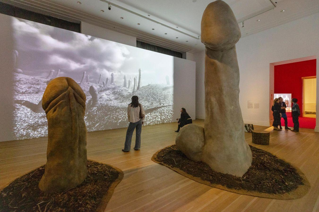 Menschen stehen in einem Ausstellungsraum, in dem zwei zwei Penis-Skulpturen zu sehen sind. Im Hintergrund sind auf einer Leinwand Penisse als Wald abgebildet.