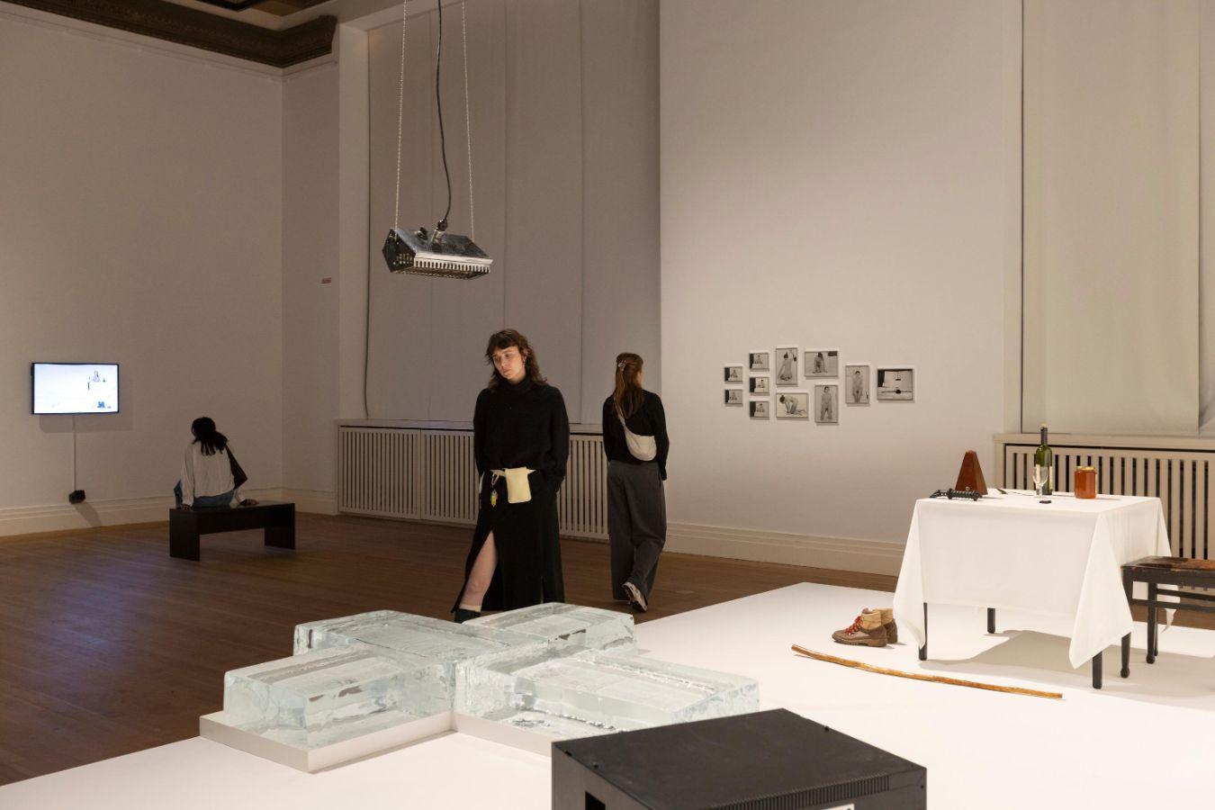 In einem Ausstellungsraum liegt auf einer weißen Erhebung ein Kreuz aus Eis, darüber hängt eine Wärmelampe. Rechts davon steht ein Tisch mit einer Weinflasche, einem Honigsglas und einem Metronom. Davor stehen Schuhe. Im Raum laufen Menschen.