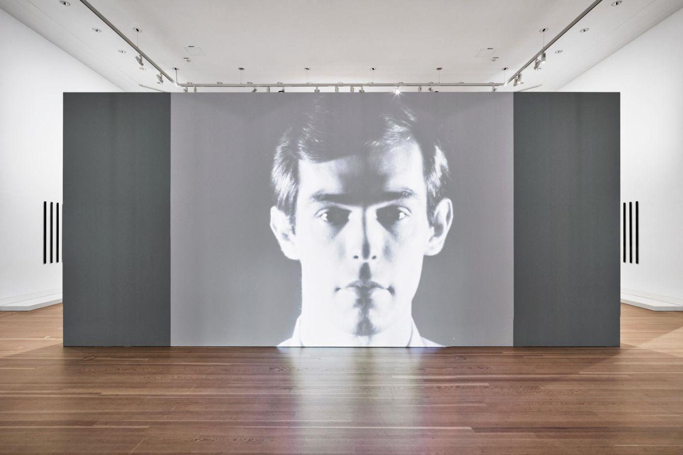 Aufnahme von einer großen Schwarz-Weiß-Projektion eines Gesichts von einer männlich gelesenen Person.