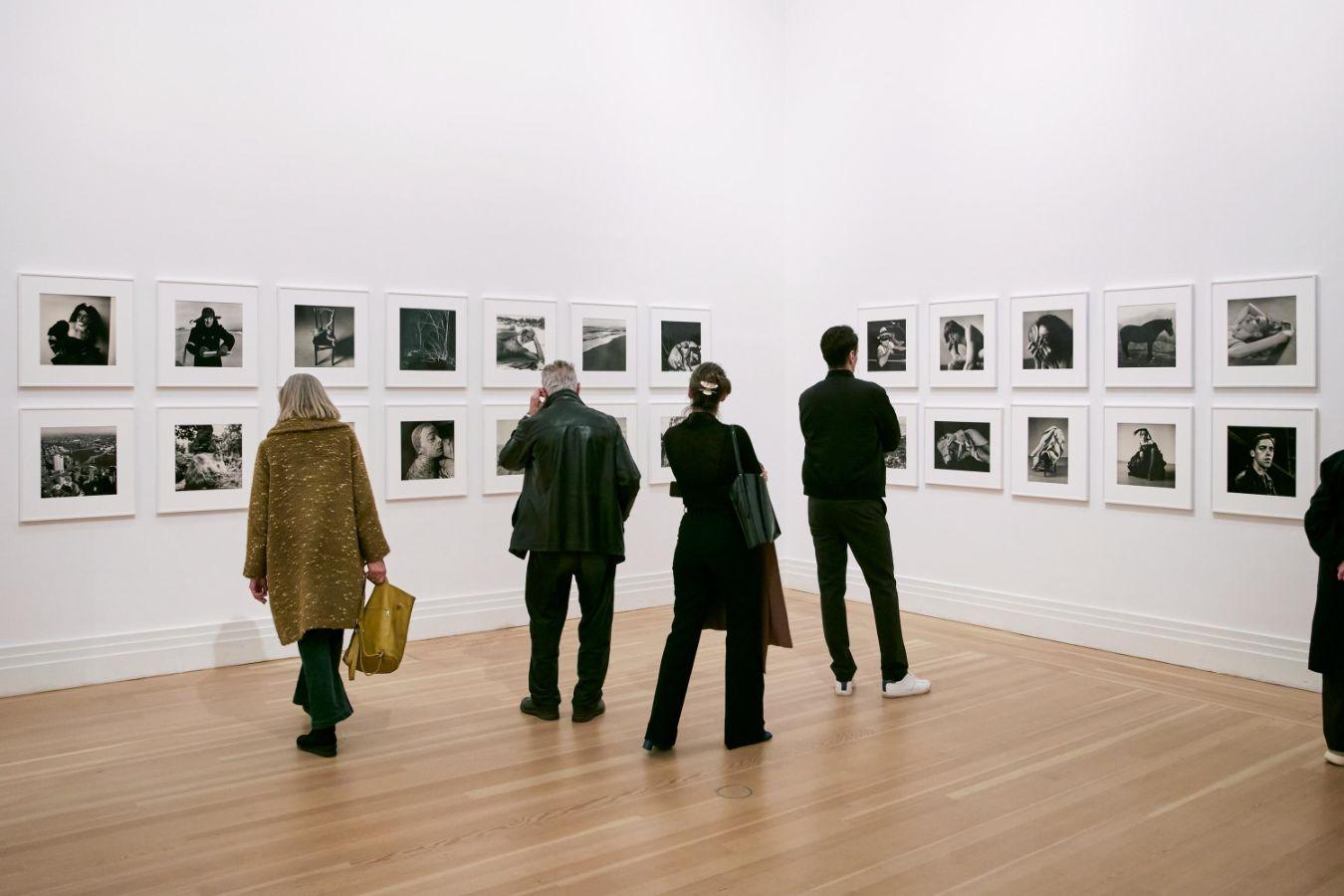 Menschen, die in einem weißen Raum vor einer Wand mit vielen quadratisch angeordneten Schwarz-Weiß-Fotografien stehen und sie betrachten.