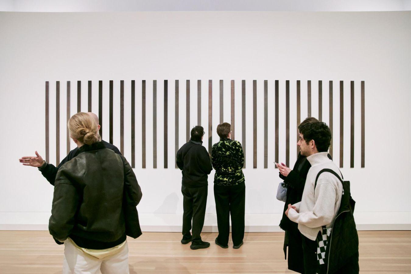 People standing in a white room in front of an artwork made of many elongated silver rods, observing it.