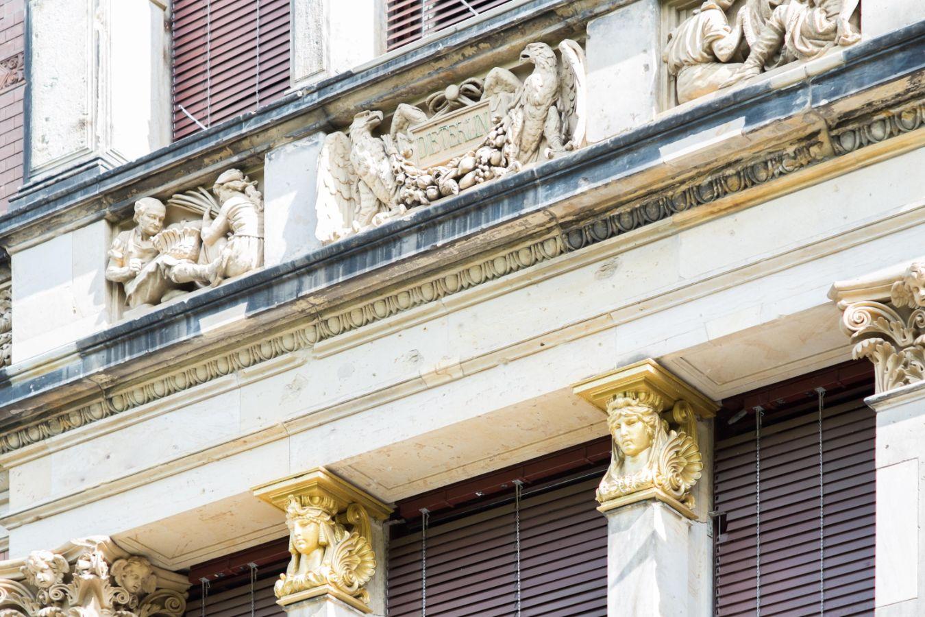 Aufnahme der Fassade eines Gebäudes im Stil der italienischen Renaissance. Neben dem Fries und Säulen mit Köpfen als Kapitell sind braune Außenjalousien zu sehen.