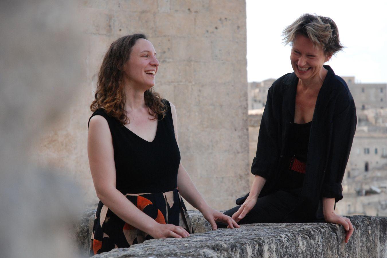 Silvia Tarozzi und Deborah Walker stehen an einem Geländer aus Stein und lachen.