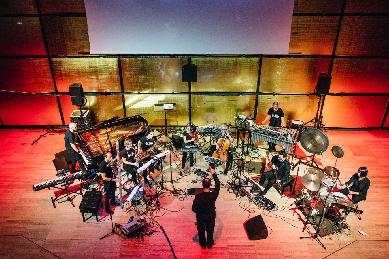 Bird's eye view of a person conducting and an orchestra of nine musicians. A video work can be seen on a screen in the background. The room is lit up in shades of red and yellow.