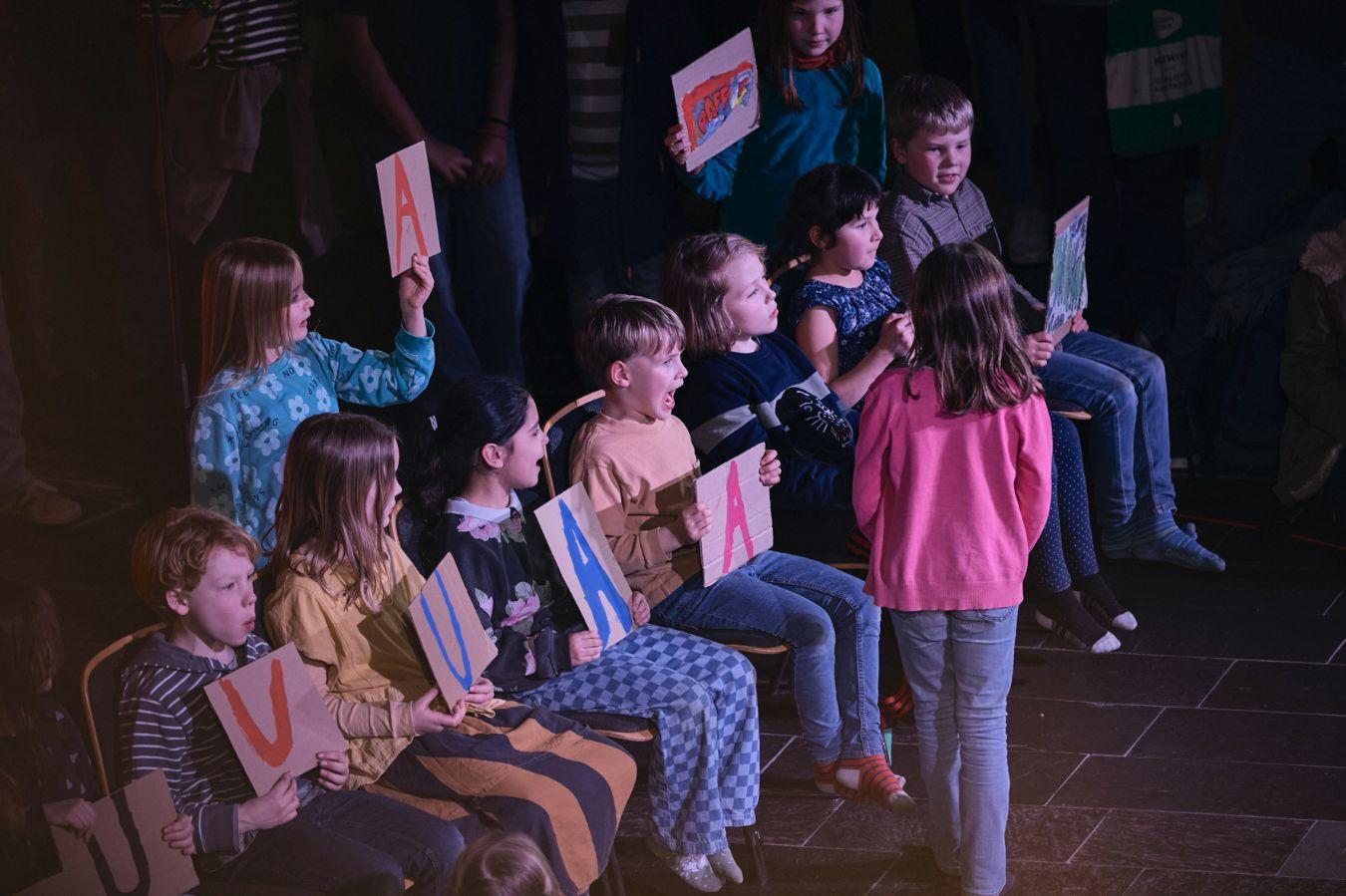 Mehrere Kinder sitzen auf Stühlen, drei Kinder stehen. Die meisten von ihnen halten Karton-Schilder hoch, mit gemalten Bildern oder den Buchstaben „A“ oder „U“. Einige Münder der Kinder sind geöffnet und formen Vokale.