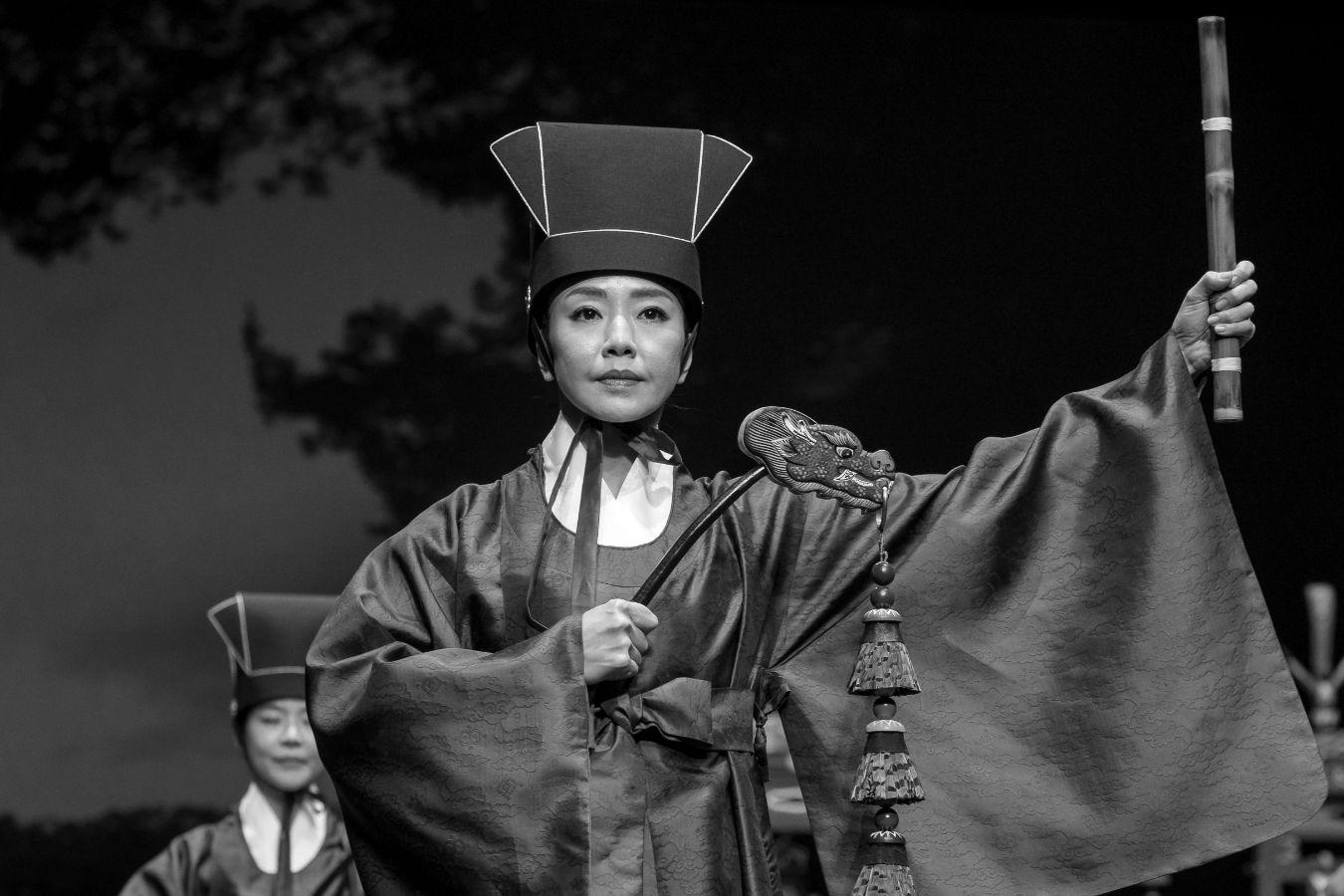 Eine Frau in rituellem Gewand hält Stock und Glockenstab in die Höhe.