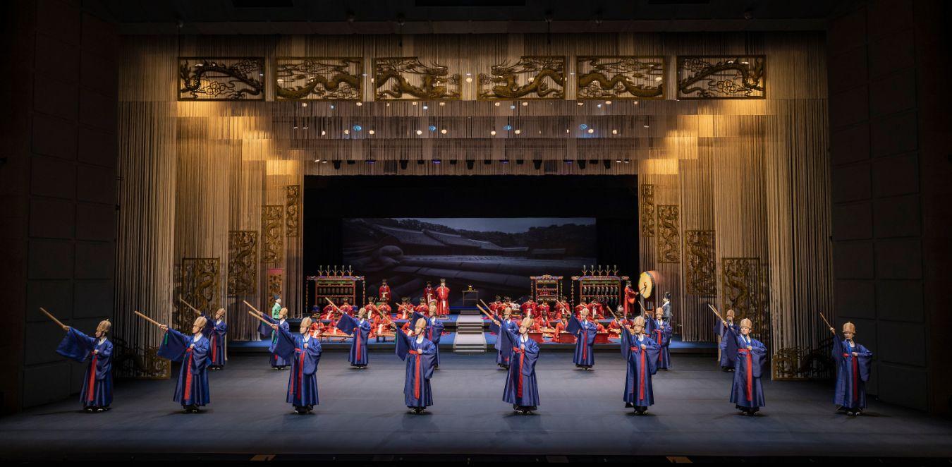 Men in ritual blue robes hold wooden swords in their hands, behind them is an orchestra in ritual red robes.