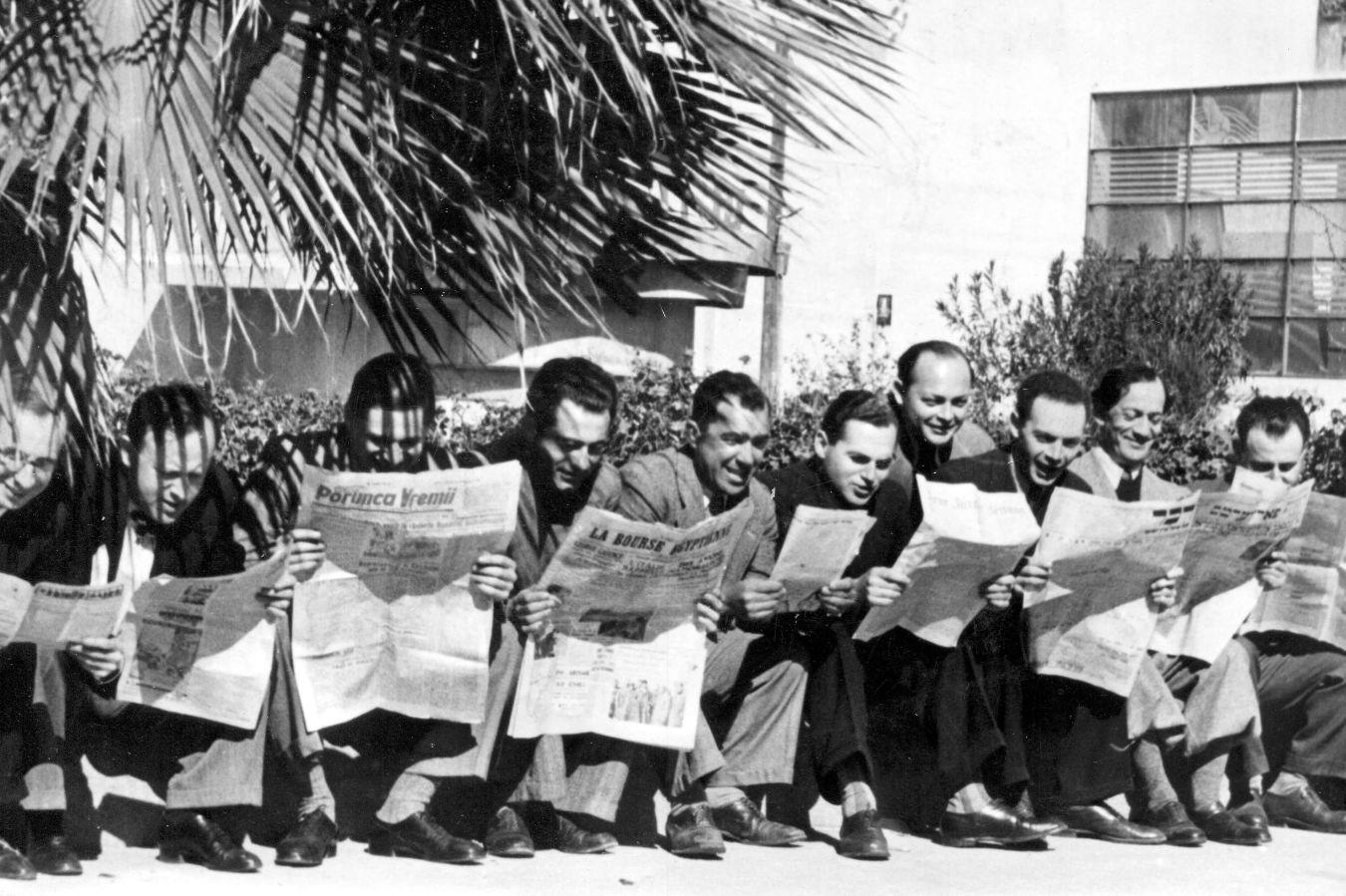 Men sit on a low bench under palm trees and read the newspaper