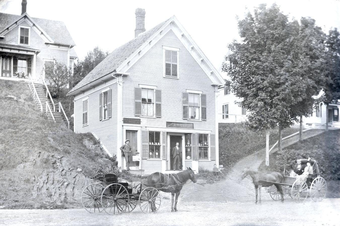 Street with house with shop front, in front of it two men and two carts with horses
