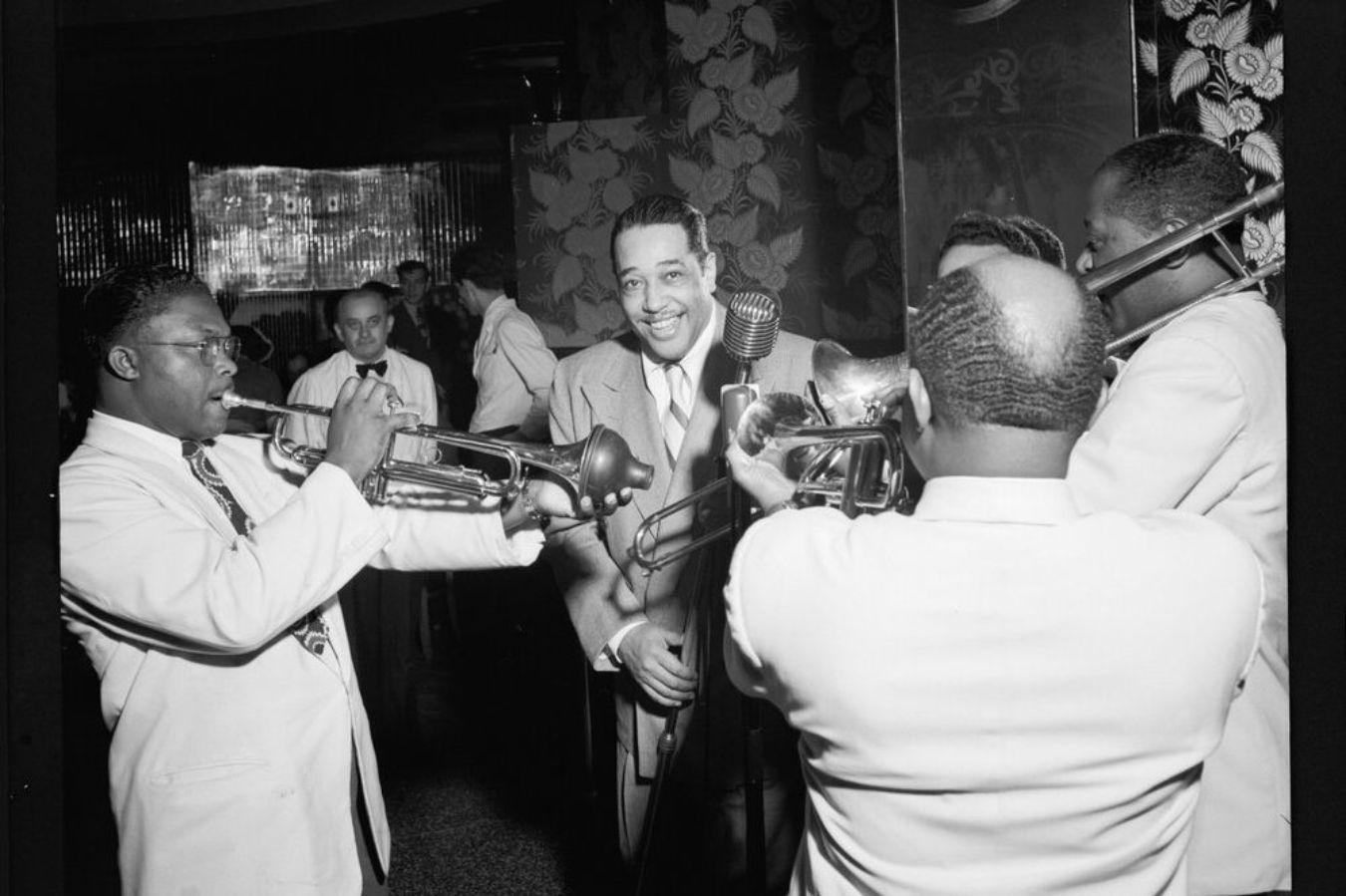 Vier Personen mit Instrumenten spielen auf einen frontal den Betrachtenden zugewandten Mann mit Mikrofon zu.