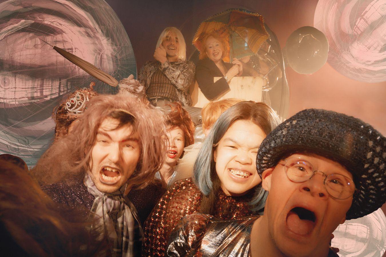 The ensemble of the piece pulls a covered wagon towards the viewer. Shiny metallic spheres reflecting the surroundings float in the background.