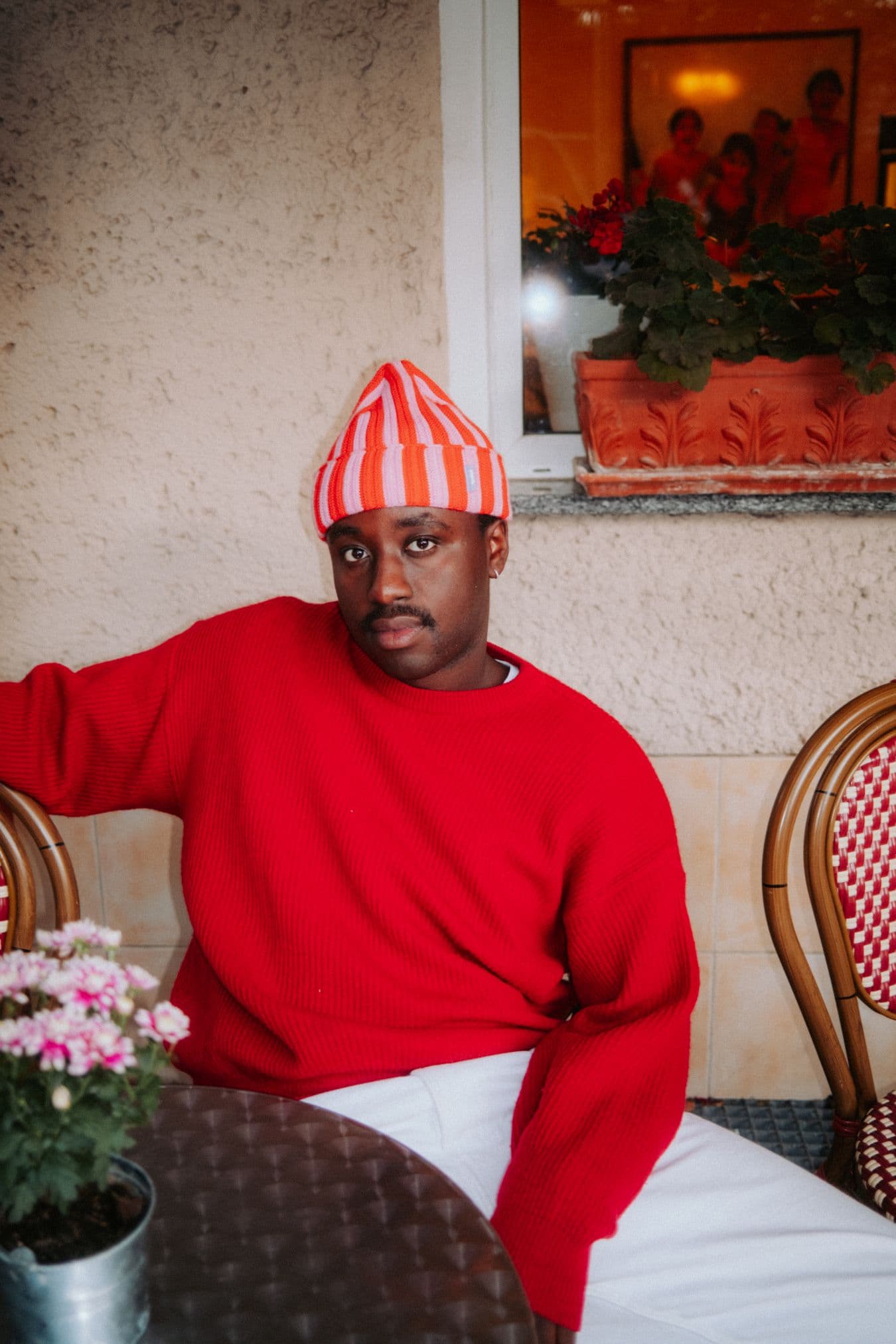 Männliche gelesene Person mit rotem Pullover und gestreifter Mütze, sitzt an einem Tisch, Blumen im Vordergrund.