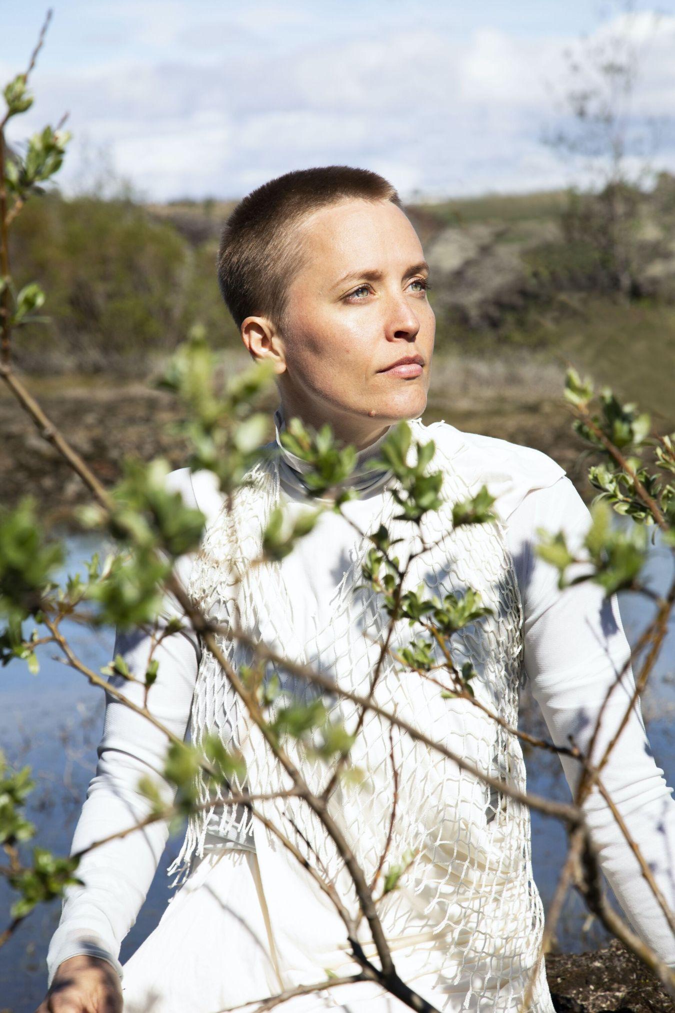A person with short hair and a white dress stands in front of a countryside background. In the foreground, branches with green buds cover part of the person