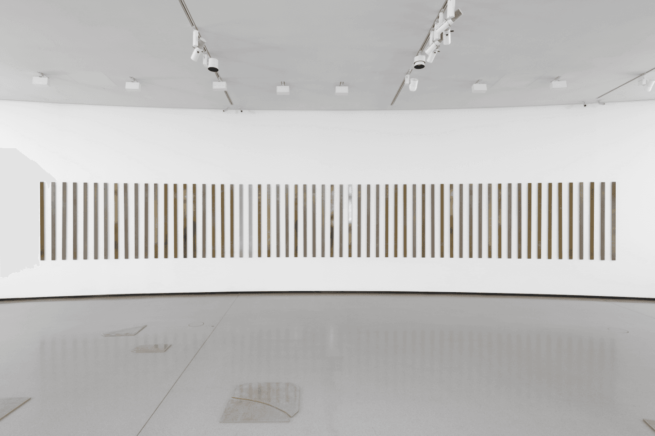 Schmale Fotografien in Braun- und Grautönen hängen nebeneinander an einer weißen Wand in einem Ausstellungsraum.