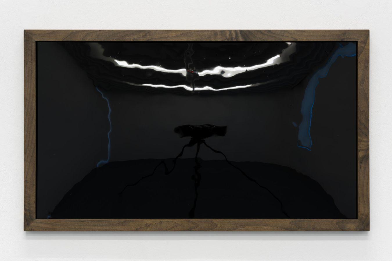 Ein glänzendes schwarzes Rechteck ist in einen dunklen Holzrahmen gefasst und horizontal an einer weißen Wand angebracht. Die reflektierende Oberfläche spiegelt das Licht des Raumes und macht die Umgebung schemenhaft sichtbar. Das Kunstwerk ist frontal aufgenommen.