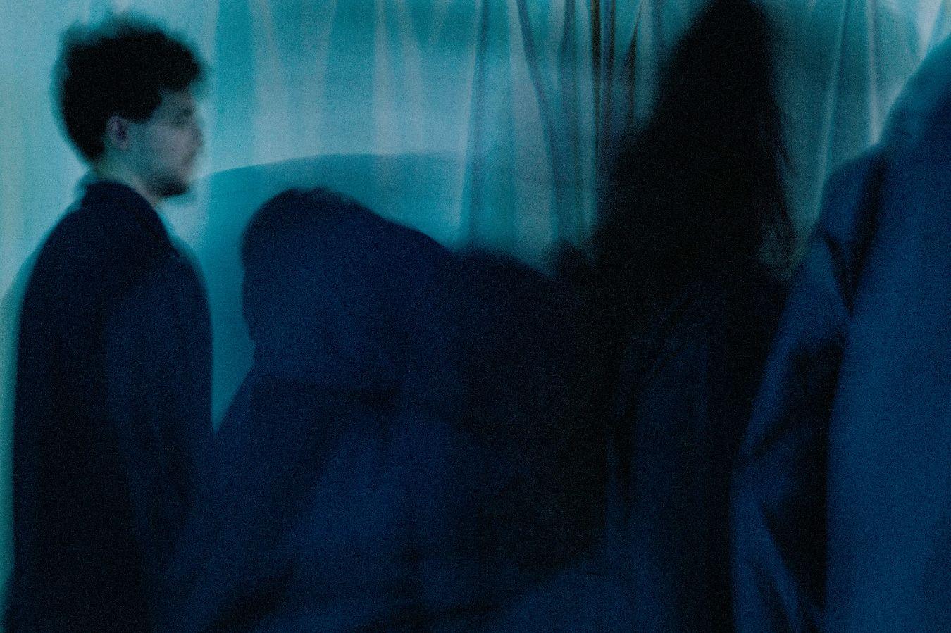 A blurred, male read person and the shadows of other persons in front of a blue curtain