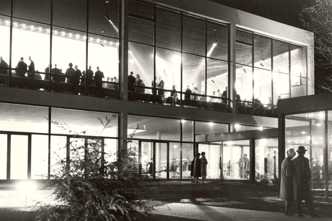 A house with a large glass façade, brightly lit inside, the upper floor full of people.