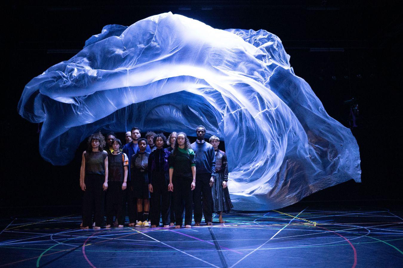 A man jumps into the air. Behind him, in the cone of light, the floor of a stage with colourful circles. To the right, a group of other dancers in motion.