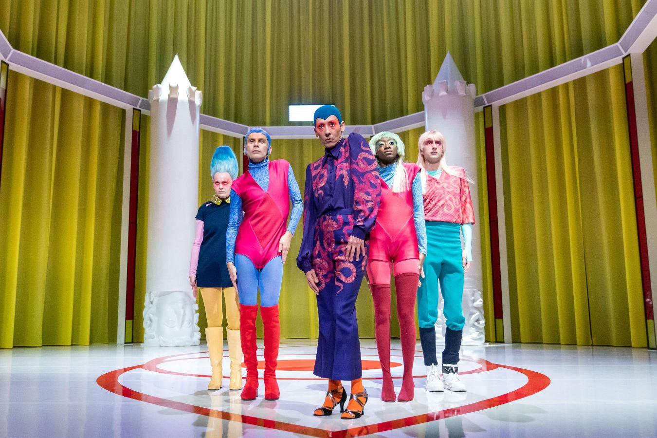 Five people stand on a stage, behind them are two large white pillars resembling rockets, poles or castle towers.