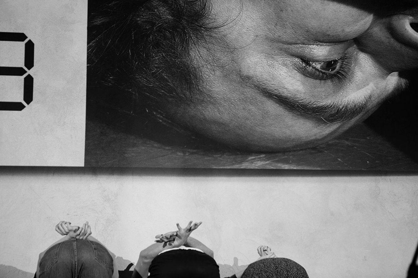 Drei Personen befinden sich mit den Händen auf dem Rücken auf einer Bühne, mit dem Rücken zum Publikum. Auf einer Leinwand über ihnen ist das Gesicht eines Mannes in Nahaufnahme zu sehen.