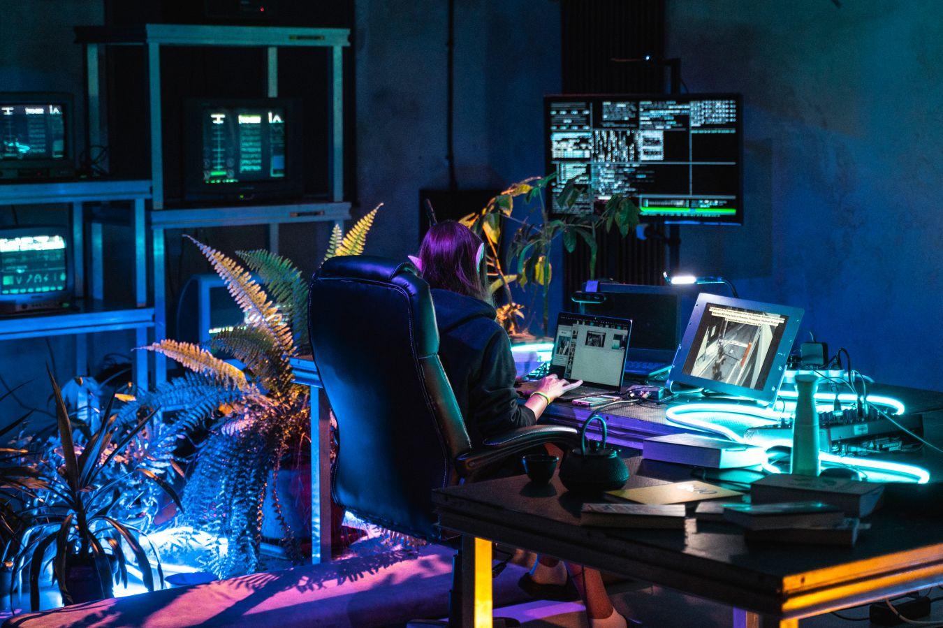 Eine Frau mit Elfenohren sitzt mit dem Rücken zur Kamera an einem Schreibtisch mit zwei Bildschirmen. Um sie herum sind Pflanzen und viel Technik.