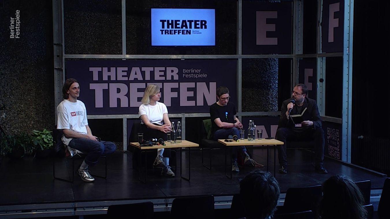 Zwei Frauen und zwei Männer sitzen um einen kleinen Tisch herum, hinter ihnen der Schriftzug Theatertreffen. Der Mann ganz rechts hat ein Mikrofon in der Hand.