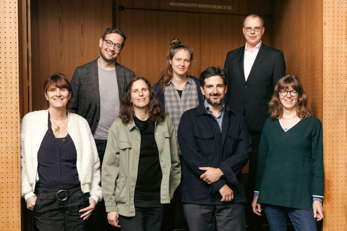 Die sieben Theatertreffen-Jurymitglieder stehen vor einer Saaltür in einem Theater und schauen in die Kamera.