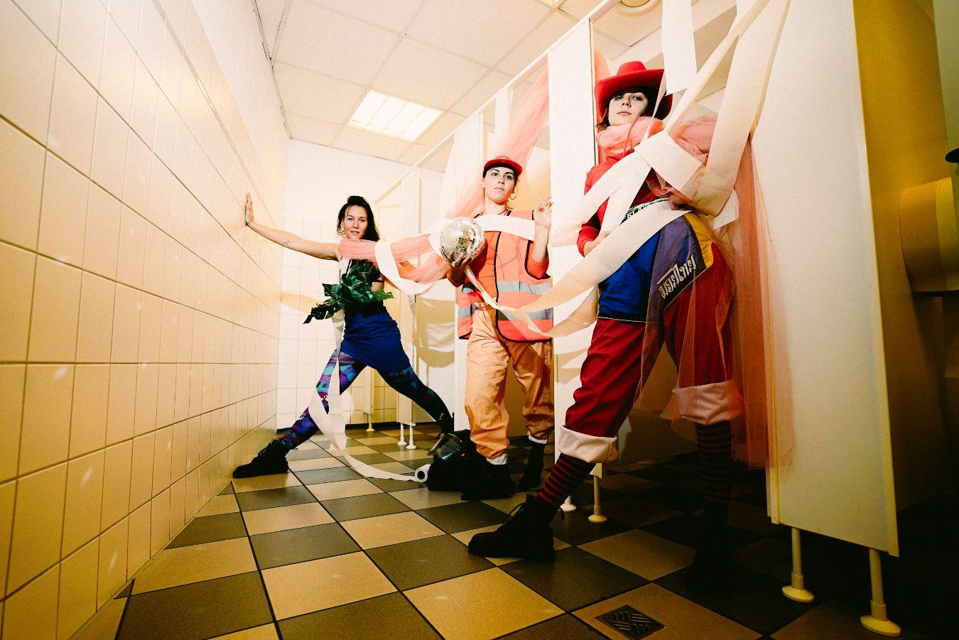 Three women in colourful costumes in a bright toilet room, with a disco ball and draped toilet paper in their hands.