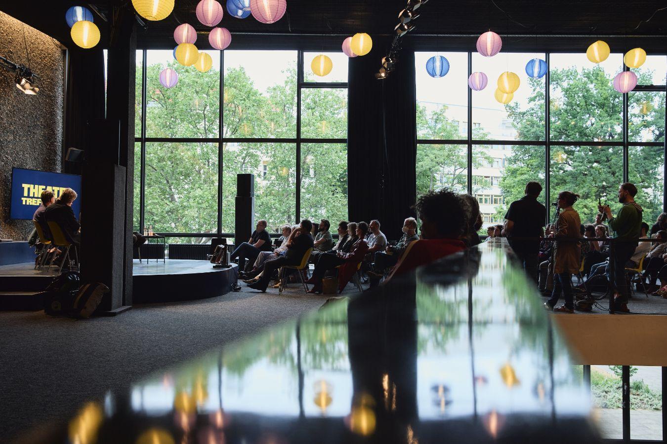 Seitlicher Blick auf das Publikumsgespräch im Oberen Foyer. Links die Bühne, in der Mitte und rechts das Publikum, im Vordergrund unscharf ein Geländer, in dem sich die Lampions an der Decke und die Fester im Hintergrund spiegeln.