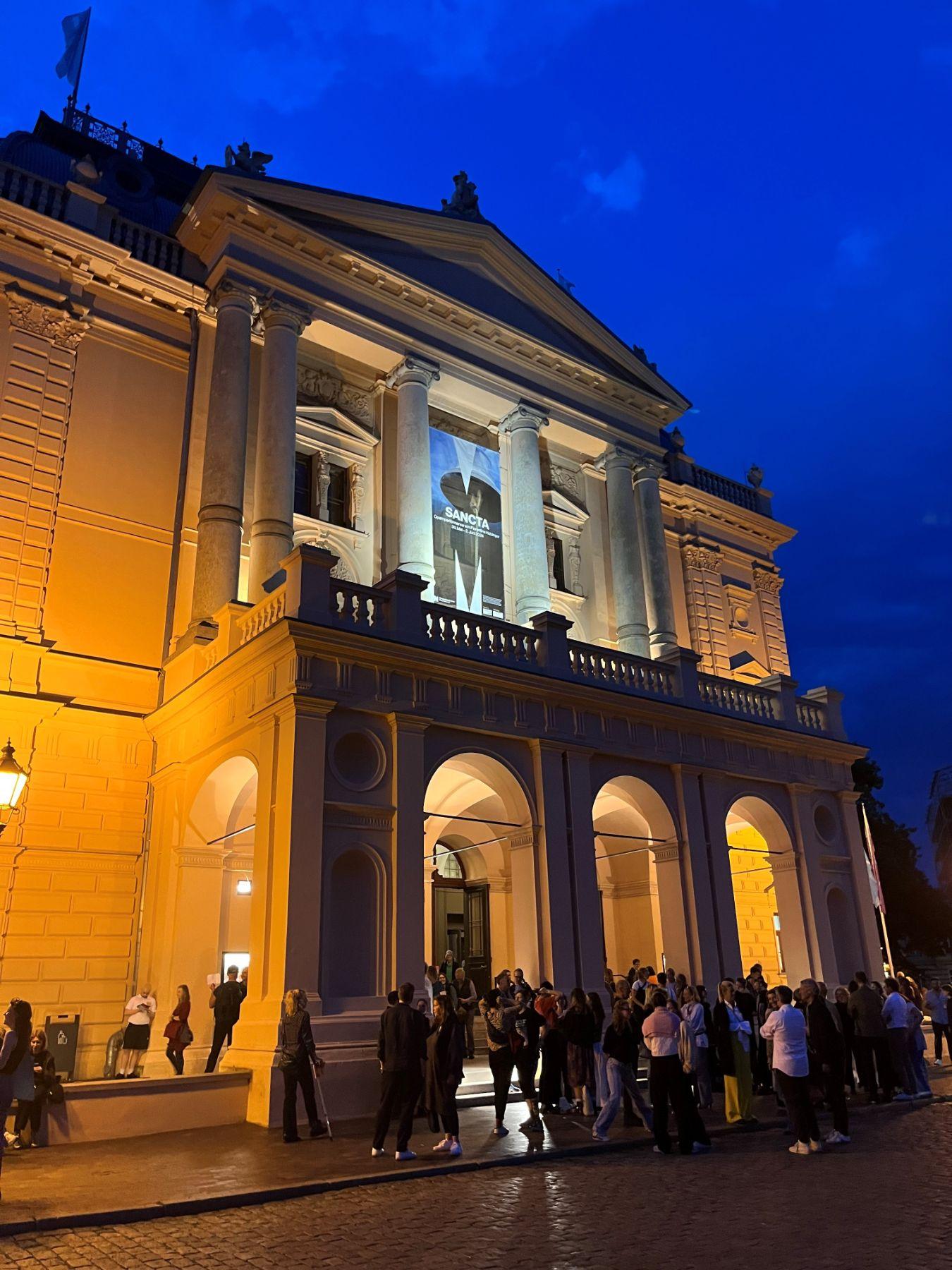 Audience gathers in front of the brightly lit Mecklenburg State Theatre in Schwerin for a “SANCTA” performance