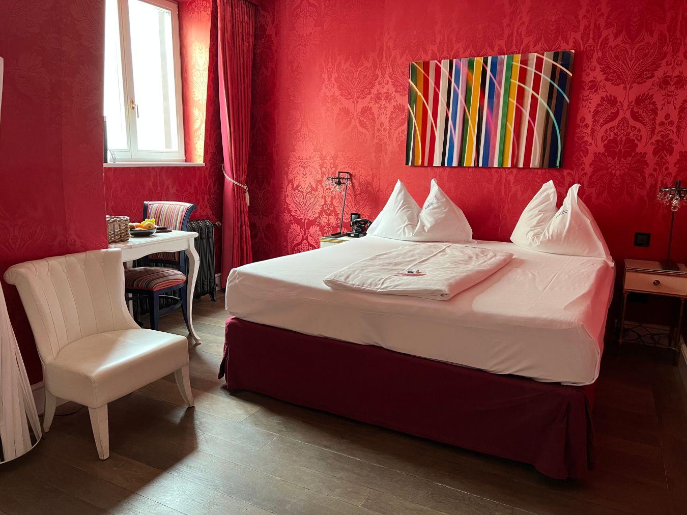 A hotel room with red damask wallpaper, white furniture and a modern picture with tasteful colourful vertical stripes