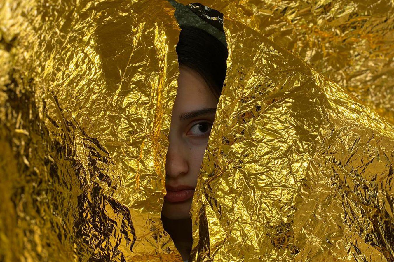 A young woman looks through a tear in a golden foil.