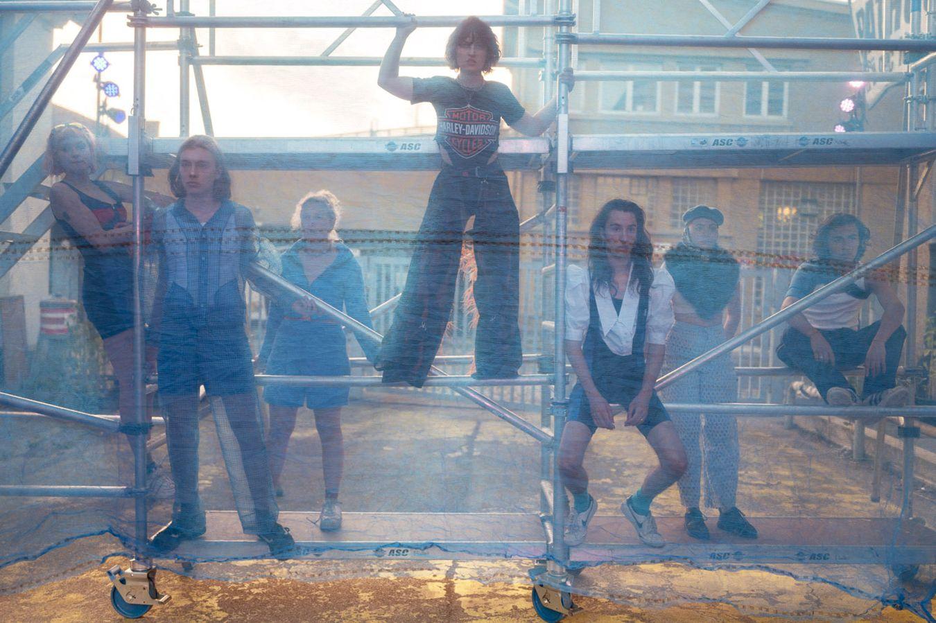 Seven actors are on a scaffolding with different postures and look through a net.
