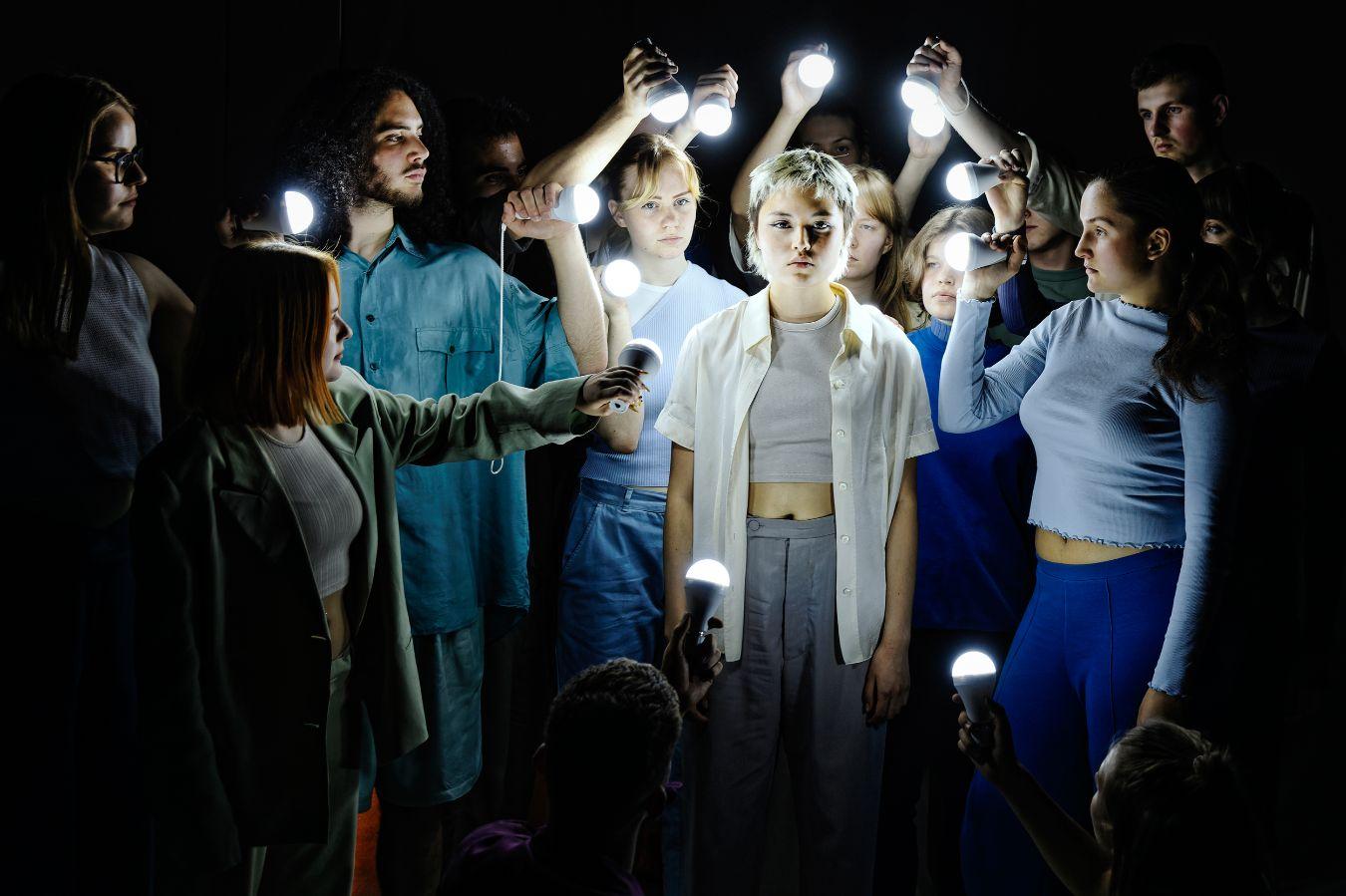 Eine kurzhaarige junge Darstellerin mit ernster Mimik wird von einer Gruppe junger Schauspieler*innen umringt, die sie mit LED-Birnen beleuchten.