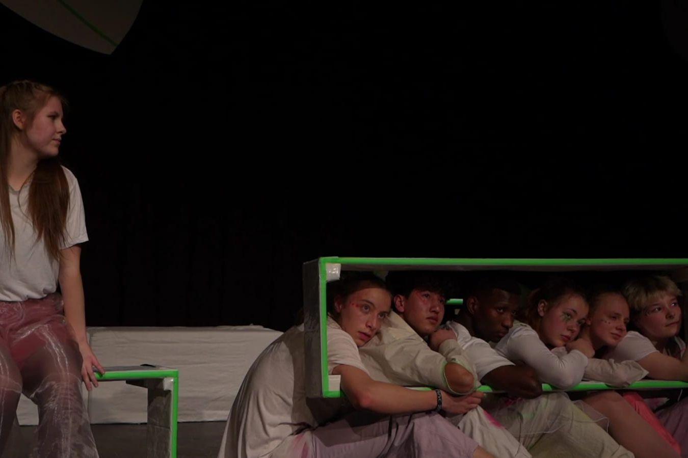 Six actors sit side by side on a stage, looking through a green, rectangular, window-like box that frames their heads and in which they rest their right arm.