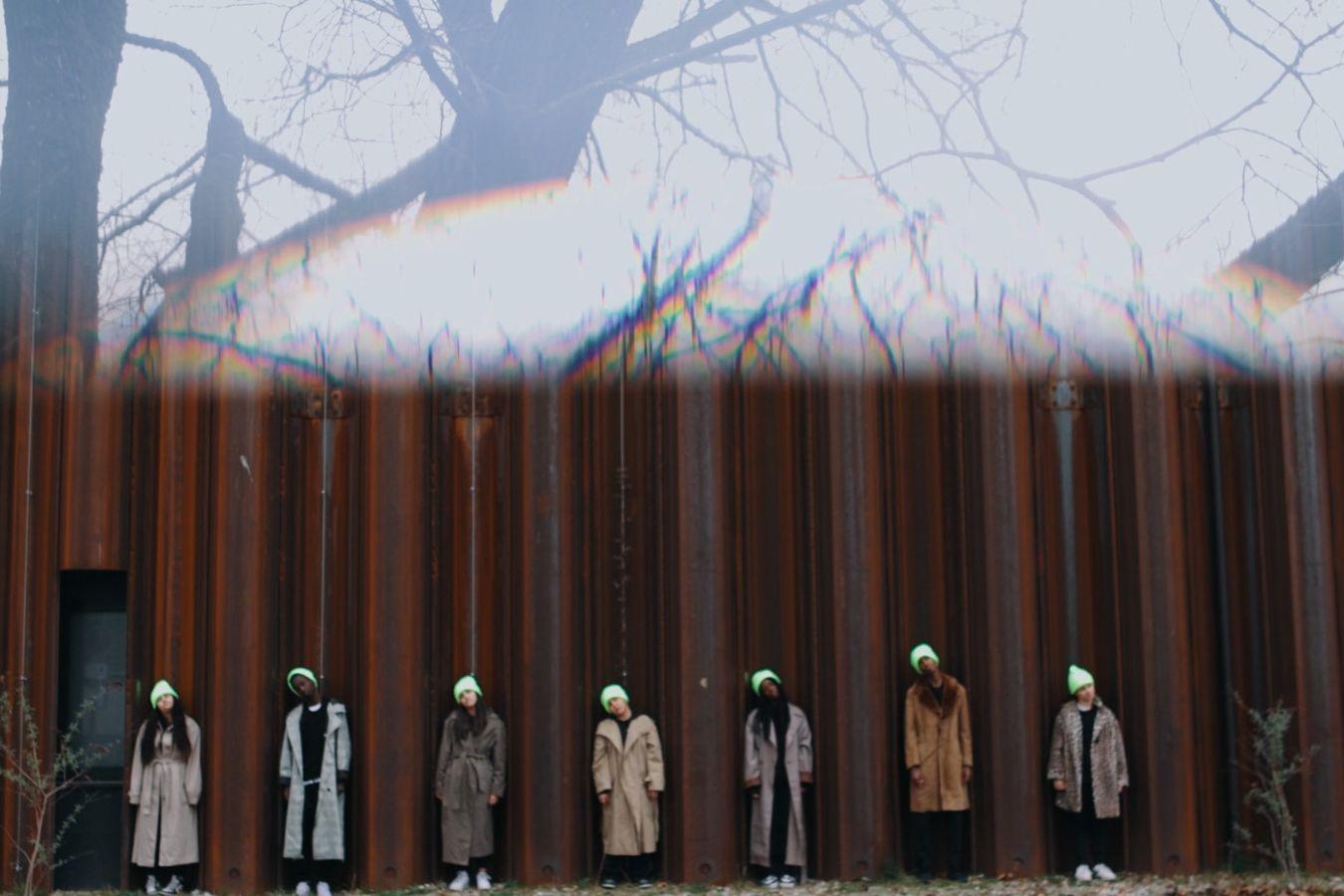 Bottom half of the picture: Seven young people in coats and neon yellow caps stand in front of a rust-coloured building. Each person is standing between a pillar. They all tilt their heads to the left and look forwards. Upper half of the picture: We see bare trees against a white background. This part of the picture is blurred