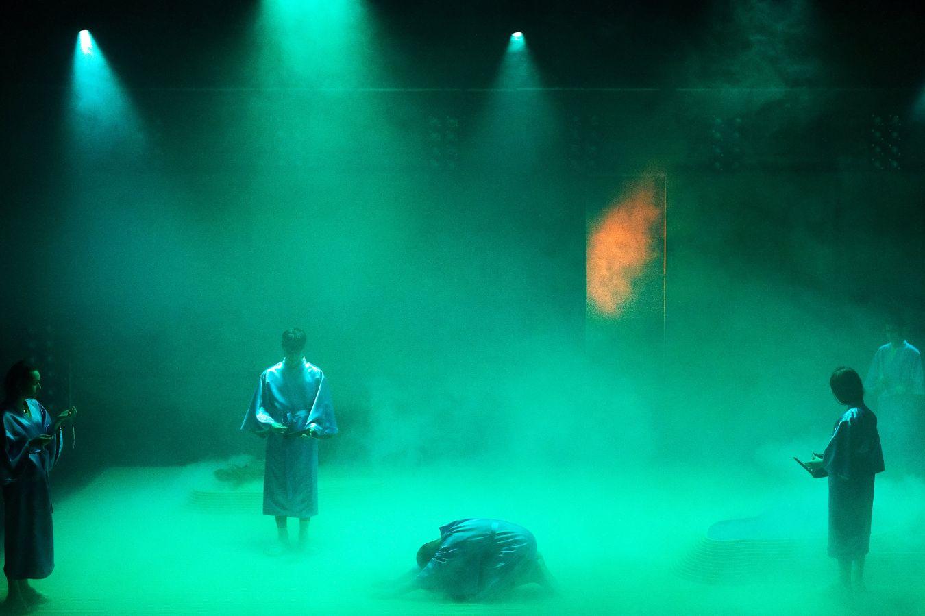 Four people stand on the stage in a green mist. Another person bows low in the centre of the stage. They are wearing blue robes. An orange glow can be seen in the middle of the fog.