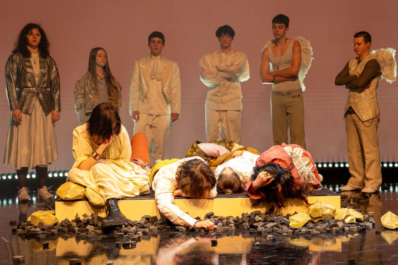Several people are lying on a mattress in the centre of the stage. There are stones and coal around the mattress. In the background, other people are standing in a semi-circle around them, two of them have angel wings.