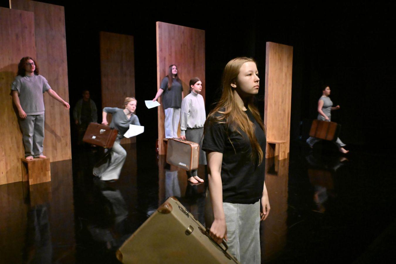 Six people are depicted on the stage set. They are all wearing gray, white, and black clothing and are distributed across the stage. Both their body posture and their gaze are directed to the right. Four of them are holding suitcases, and two of the people with suitcases also appear to be running in that direction. Two people without suitcases are standing on raised platforms. In the background, there are three wooden walls.