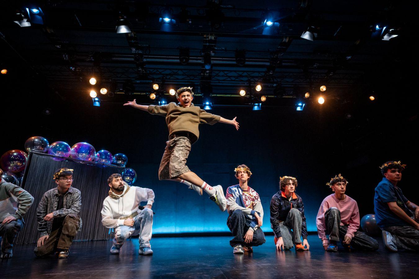 A stage photo depicting eight male-presenting individuals. Seven of them are kneeling on the floor and mostly looking straight ahead. The one in the center is jumping and is caught mid-air with arms wide open and a screaming facial expression. In the background, stage lights create a theatrical atmosphere. They are wearing typical youthful clothing and a golden laurel wreath on their head.