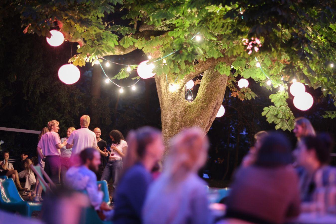 Ein großer Baum, geschmückt mit bunten Lampen, bildet den Mittelpunkt. Davor stehen und sitzen zahlreiche junge Menschen in entspannter Atmosphäre.
