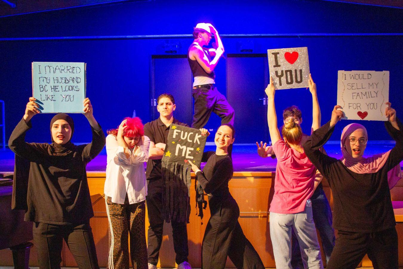 The stage photo shows seven young people. Six of them are standing in front of a stage-like raised platform, and one person is standing on it. Five of them are holding signs that read: “I married my husband because he looks like you,” “I love you,” “I would sell my family for you,” and "fuck me". The person on the platform is wearing a hat and performs a dance move reminiscent of Michael Jackson. The background is lit in blue.