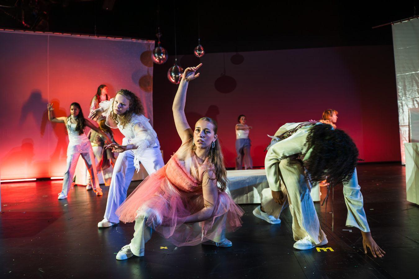 The stage photo shows approximately seven people. The right side of the image shimmers in green light, while the left side is illuminated in red. In the foreground, there is a female-presenting person wearing a pink dress who is crouching. She is looking forward but not into the camera, with her arm elegantly extended above her. To the left and right of her, but further back, there are two more people who are also holding interesting poses. This continues all the way to the very back of the stage.