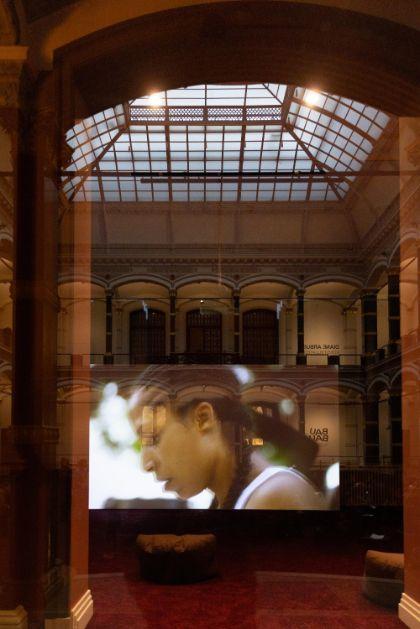 A screen is reflected in a window overlooking the historical atrium of Gropius Bau. The reflection reveals a scene from "A Plot, A Scandal" by artist Ligia Lewis. The cropped profile of Ligia Lewis is faintly visible.