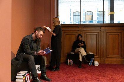 In the corner of a room with a dark red velvet carpet, there is a section with books. In this reading corner, visitors of the exhibition "Ligia Lewis: I’M NOT HERE FORRRRR…" browse through literature thematically related to the topics of the exhibition.