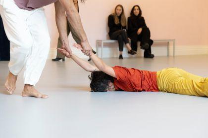 In a light pink-painted room, a live performance titled “study now steady” is taking place. Several dancers move dynamically through the space. The audience sits on simple benches, attentively observing the performance.