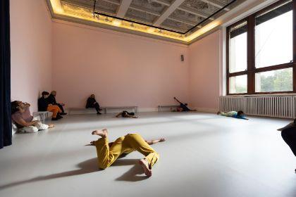 In a light pink-painted room, a live performance titled “study now steady” is taking place. Several dancers move dynamically through the space, incorporating the floor, walls, and benches into their choreography. The audience sits on simple benches, attentively observing the performance.