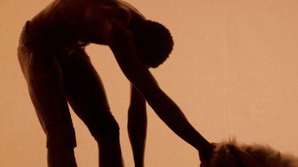 A hunched silhouette stands against a bright orange background. The head and both arms hang downward, and the person holds a fluffy object in one hand.