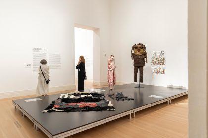 Drei Personen betrachten Kunst in einem hellen Ausstellungsraum, darunter Stoffarbeiten auf einem Podest und Kostüme.